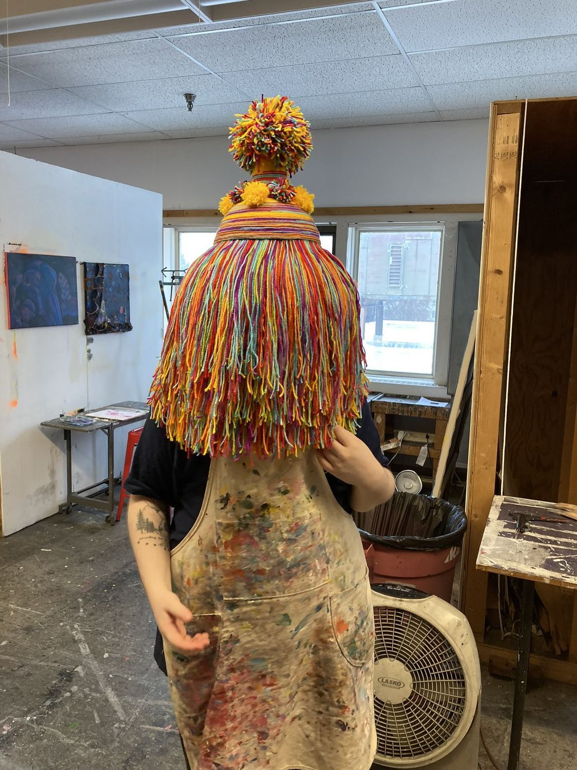 Ray A. Laventure standing in the studio wearing a hand-made yarn mask — a tall striped cone topped with pom-poms, falling to the shoulders in a curtain of rainbow fringe.