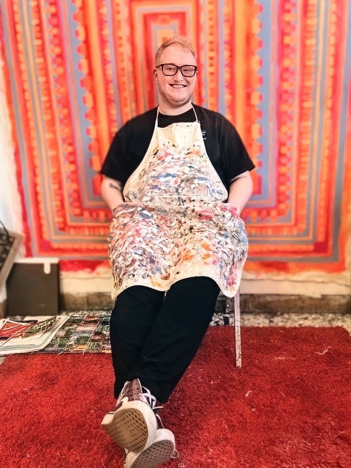 Ray A. Laventure seated and smiling in a heavily paint-spattered canvas apron, legs stretched out on a red rug, in front of a large neon orange, pink and grey concentric painting that fills the wall behind him.